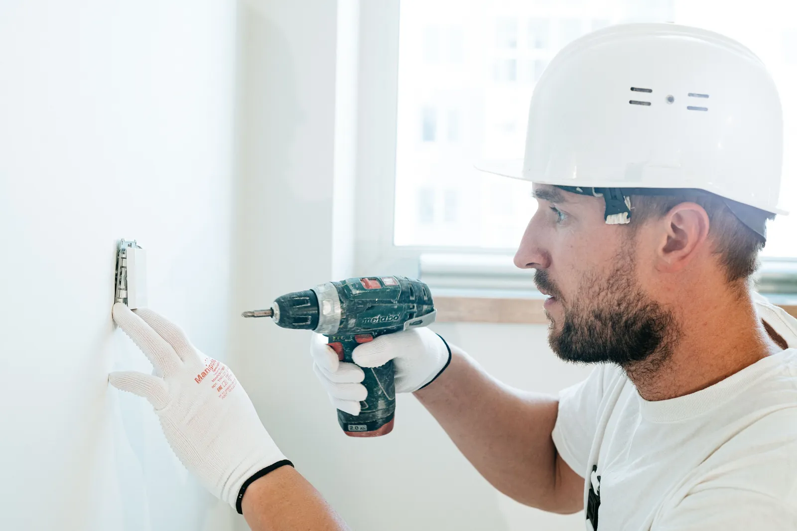 Handwerker mit Schutzhelm und Handschuhen bohrt an einer Innenwand