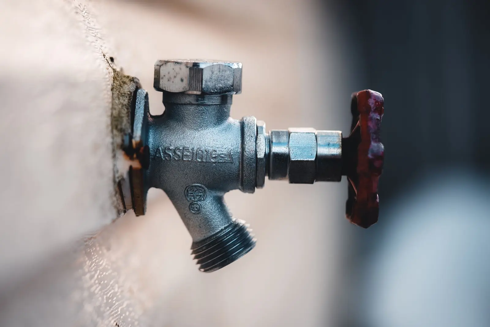 Metallischer Außenwasserhahn mit rotem Handrad an einer hellen Hauswand