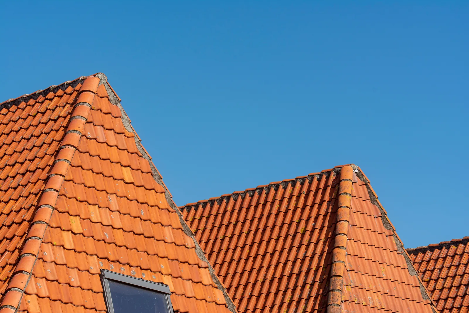 Mehrere steile Dachflächen mit roten Dachziegeln, Firsten und Dachfenster vor blauem Himmel