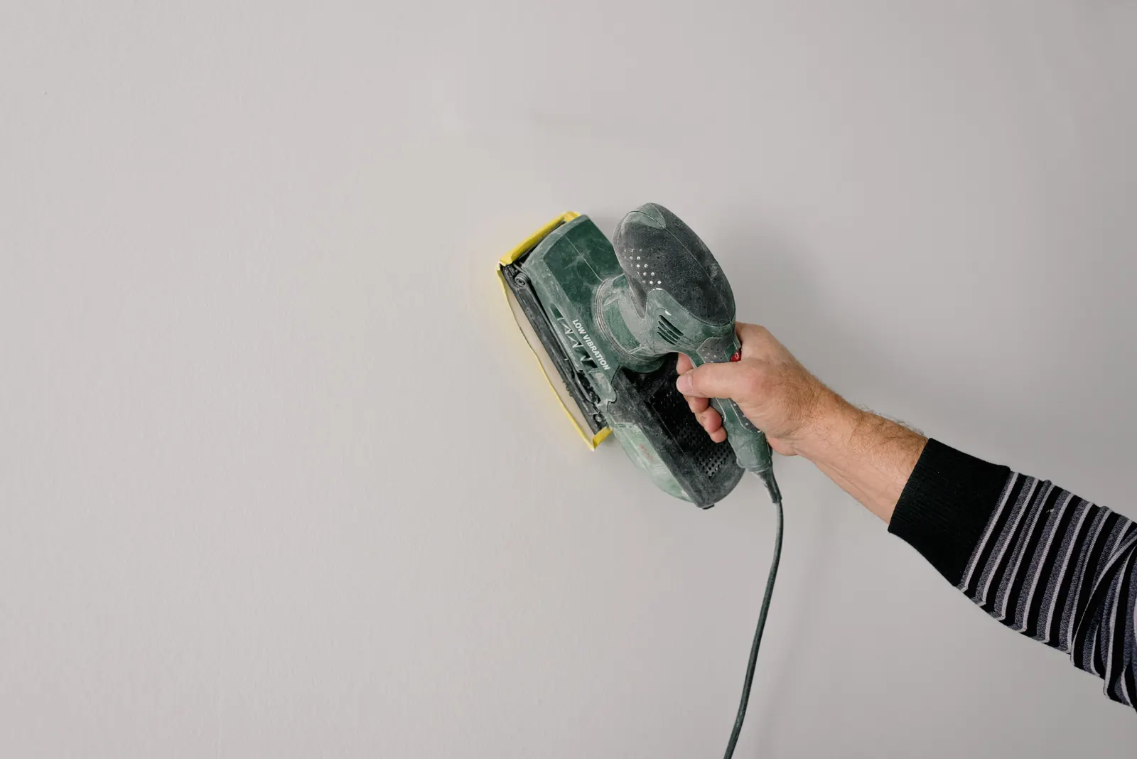 Hand führt einen elektrischen Wand- und Deckenschleifer über eine helle Innenwand
