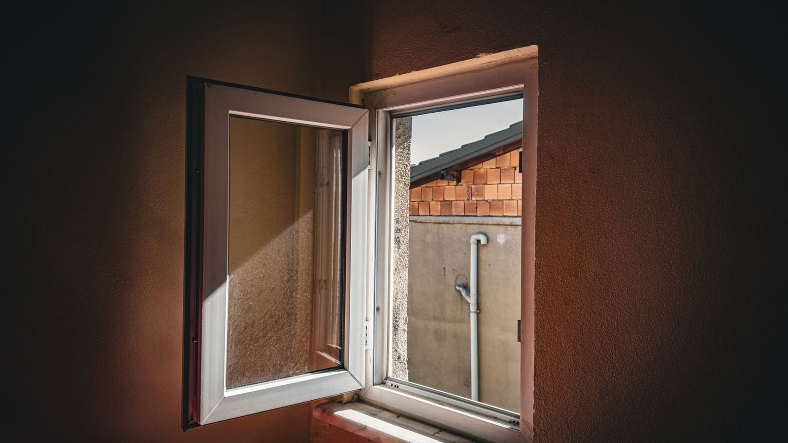 Weit geöffnetes weißes Kunststofffenster mit Blick auf eine Hofwand, davor ein dunkler Raum